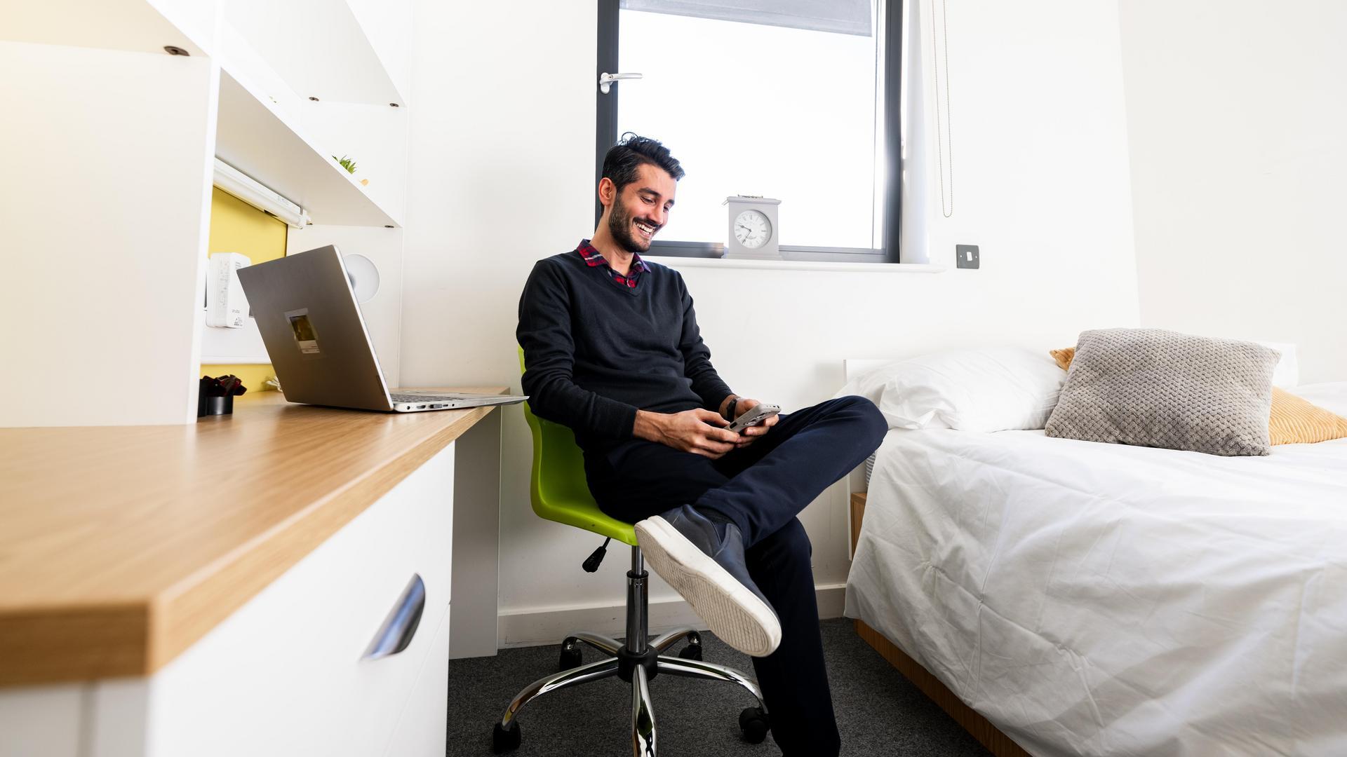 Male student in accommodation sat at desk, looking at phone