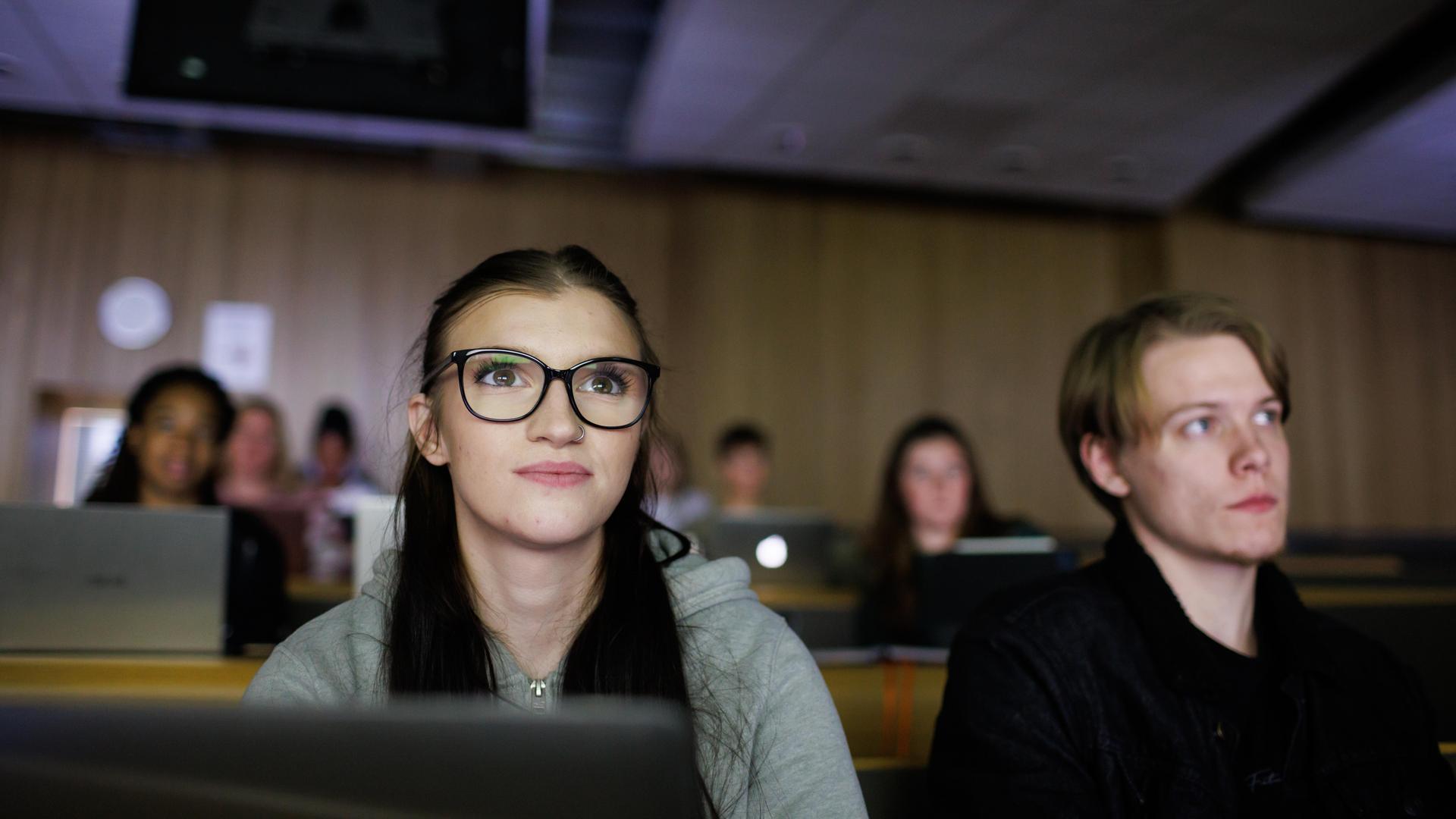 Two students are sat in a lecture theatre listening to a speaker at the front.